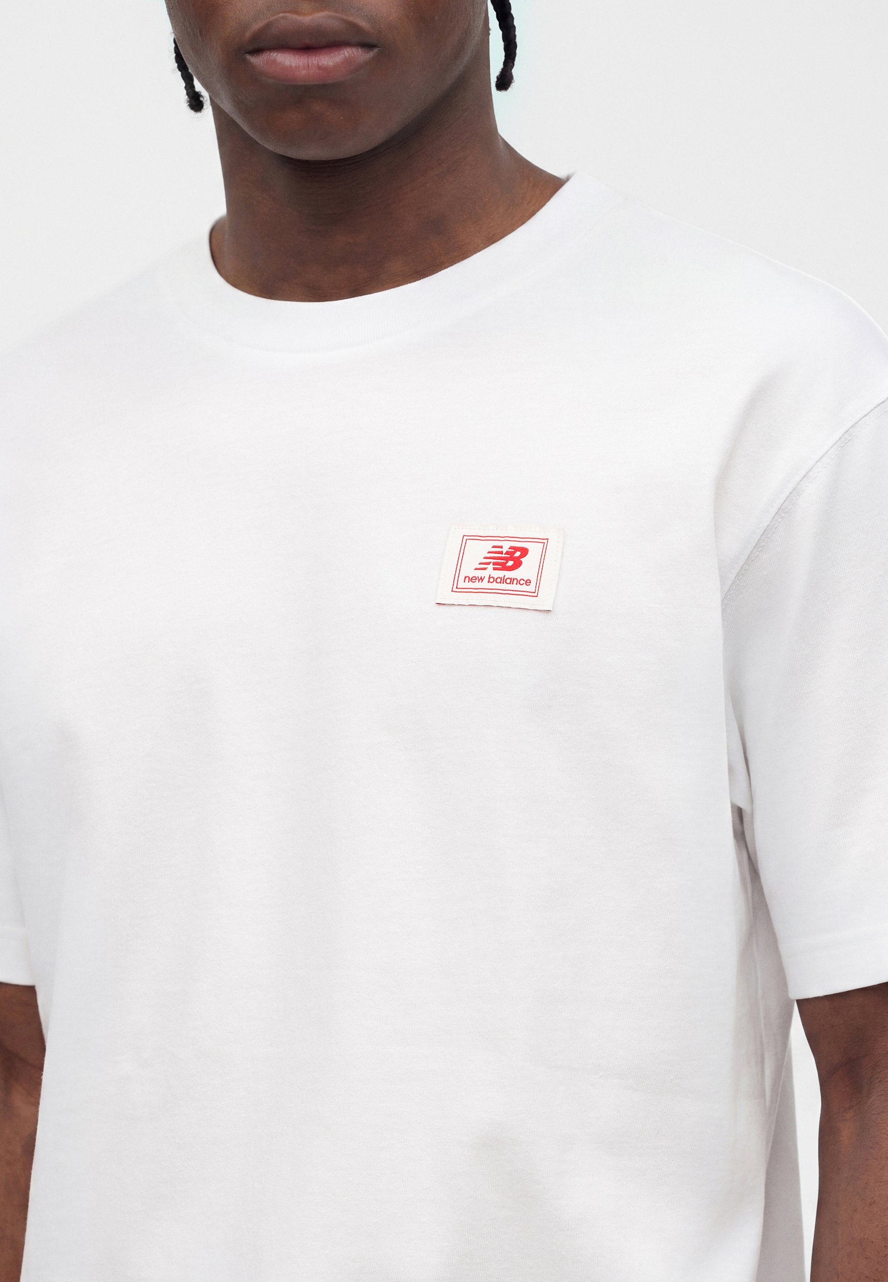 Primer plano de persona con camiseta blanca lisa con pequeño parche rojo del logo de New Balance en el pecho izquierdo.