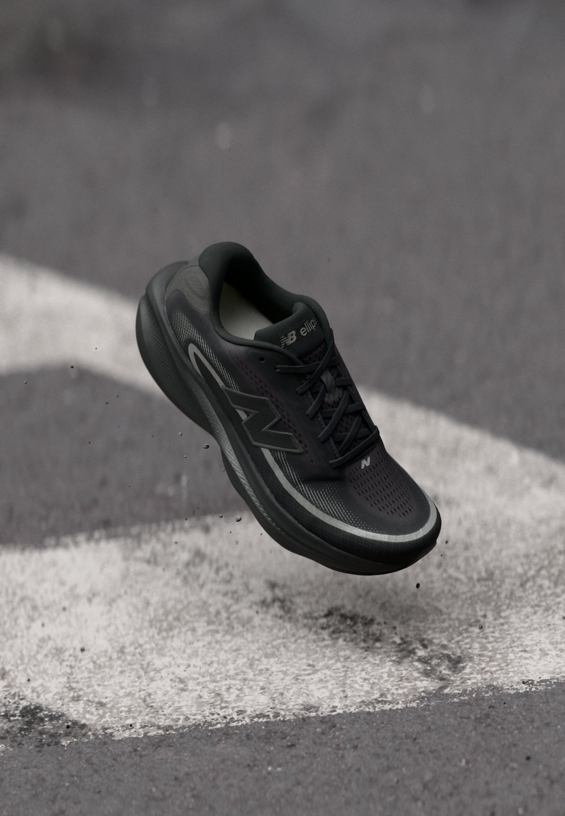 Zapatilla de running negra de New Balance suspendida sobre un asfalto con líneas blancas pintadas y pequeños escombros dispersos.