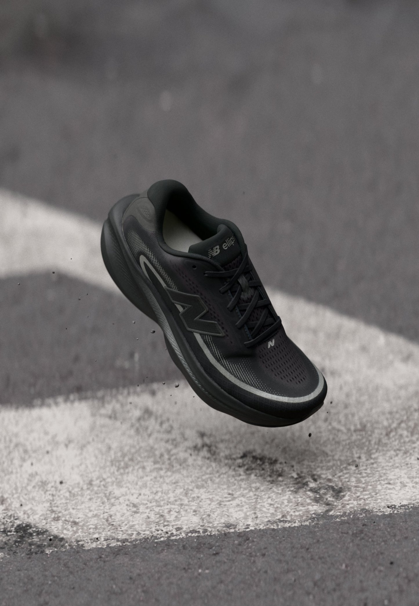 Zapatilla de running negra de New Balance suspendida sobre un asfalto con líneas blancas pintadas y pequeños escombros dispersos.