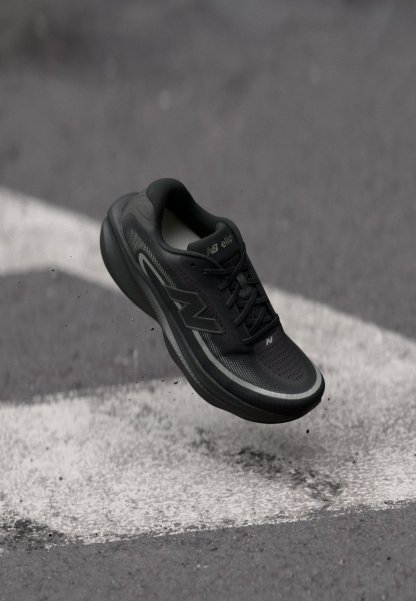 Zapatilla de running negra de New Balance suspendida sobre un asfalto con líneas blancas pintadas y pequeños escombros dispersos.