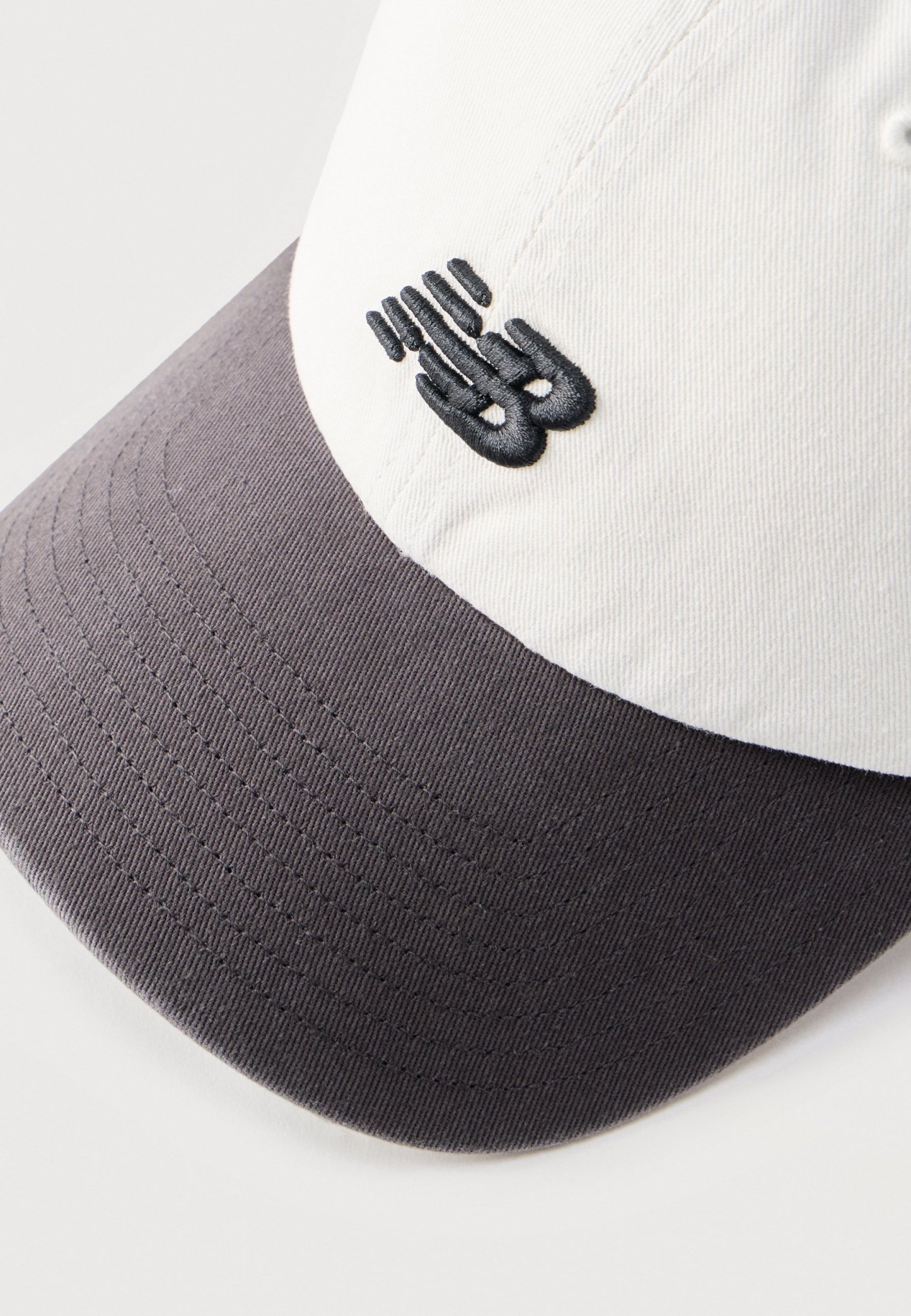 Gorra de béisbol con corona blanca y visera gris oscuro, con un pequeño logo abstracto negro bordado en el panel frontal.
