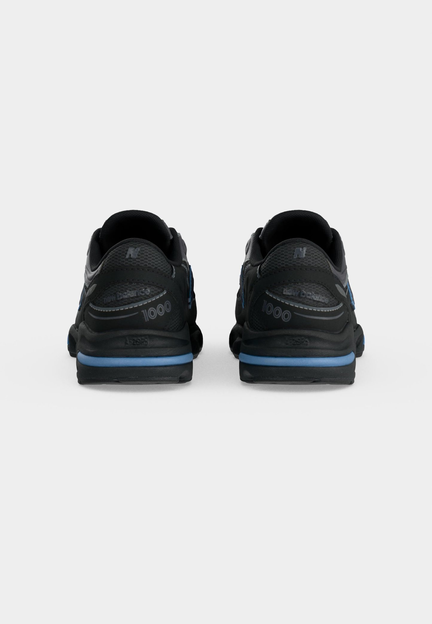 Par de zapatillas New Balance 1000 negras y azules vistas por detrás sobre un fondo claro y liso.