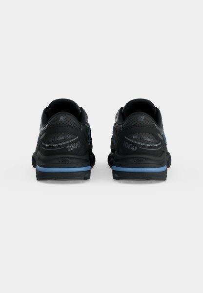 Par de zapatillas New Balance 1000 negras y azules vistas por detrás sobre un fondo claro y liso.