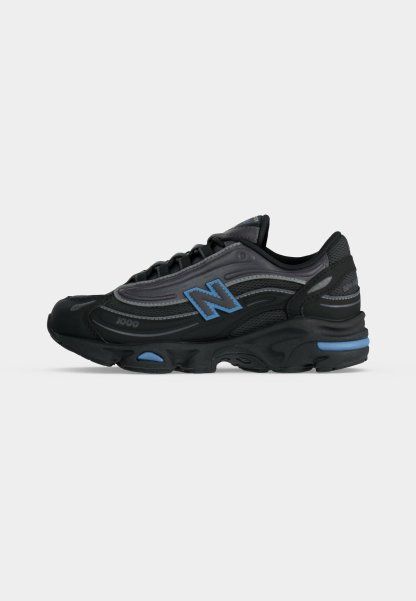 Zapatilla deportiva negra y gris oscuro con detalles en azul, diseño con cordones y un destacado logo "N" en el lateral.