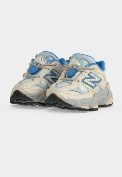 Zapatillas New Balance beige y azul para niños, con tejido de malla, suelas gruesas y tiras de velcro sobre un fondo blanco.