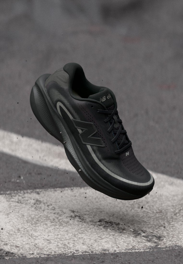 Zapatilla de correr New Balance negra en el aire sobre una superficie de asfalto con una marca blanca en la carretera, mostrando la suela y el logo lateral.