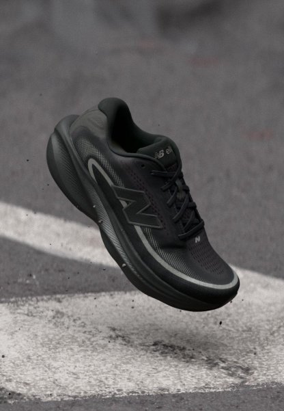 Zapatilla de correr New Balance negra en el aire sobre una superficie de asfalto con una marca blanca en la carretera, mostrando la suela y el logo lateral.
