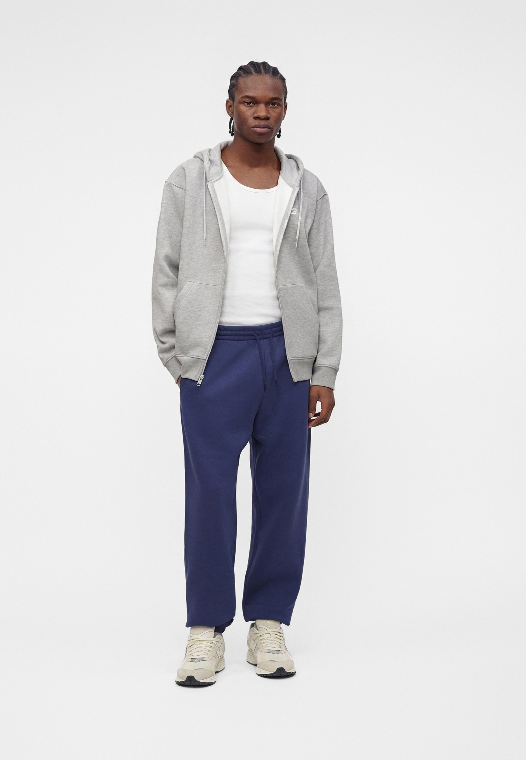 Un joven está de pie con una sudadera gris con cremallera, camiseta sin mangas blanca, pantalones deportivos azul marino y zapatillas blancas sobre un fondo blanco liso.