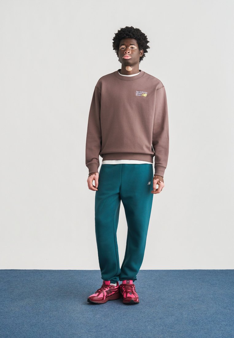 Sudadera marrón con un pequeño logo, pantalones deportivos verde azulado y zapatillas rosas. El conjunto presenta un ajuste relajado y texturas suaves.