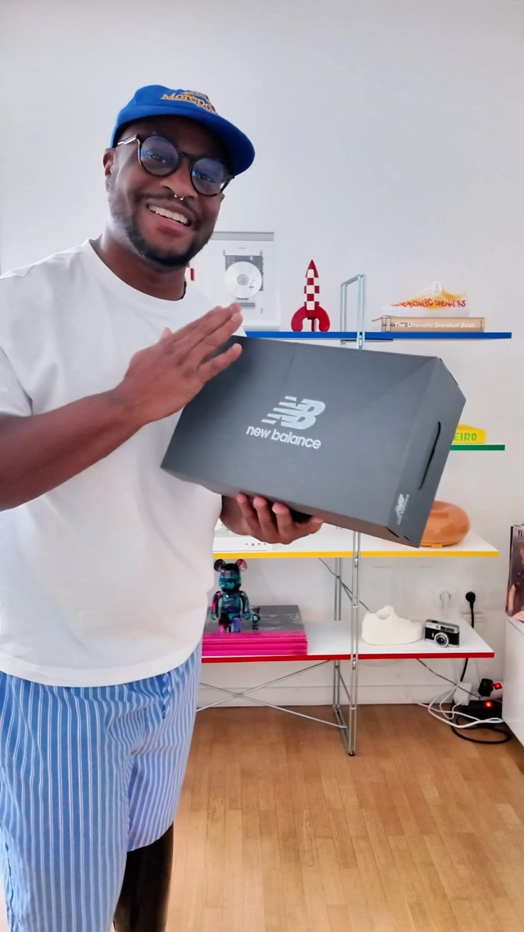 Caja de zapatos negra de New Balance con logo blanco, sostenida en las manos. Presenta una forma rectangular y tapa plana con cortes laterales para las asas.
