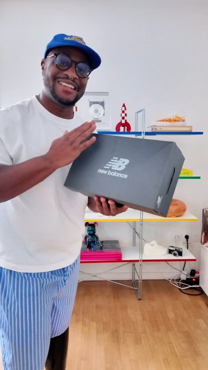 Caja de zapatos negra de New Balance con logo blanco, sostenida en las manos. Presenta una forma rectangular y tapa plana con cortes laterales para las asas.