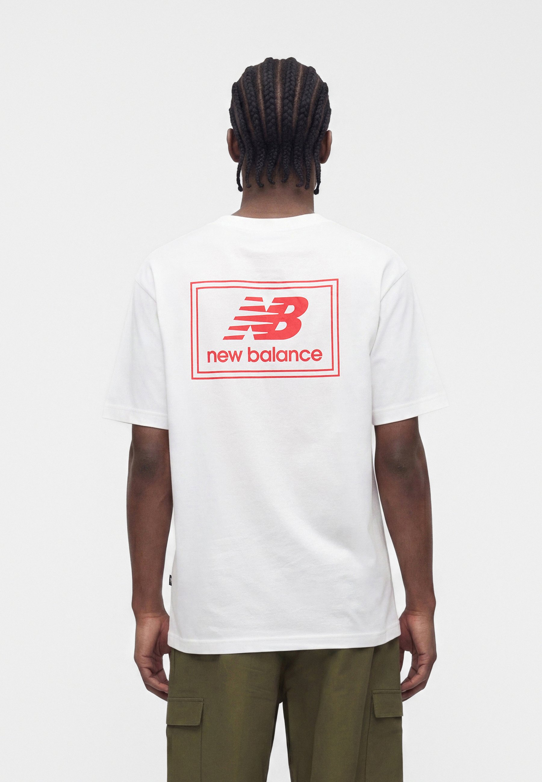 Persona con el cabello trenzado que lleva una camiseta blanca con el logo rojo "NB new balance" en la parte trasera y pantalones cargo verde oliva, de espaldas.