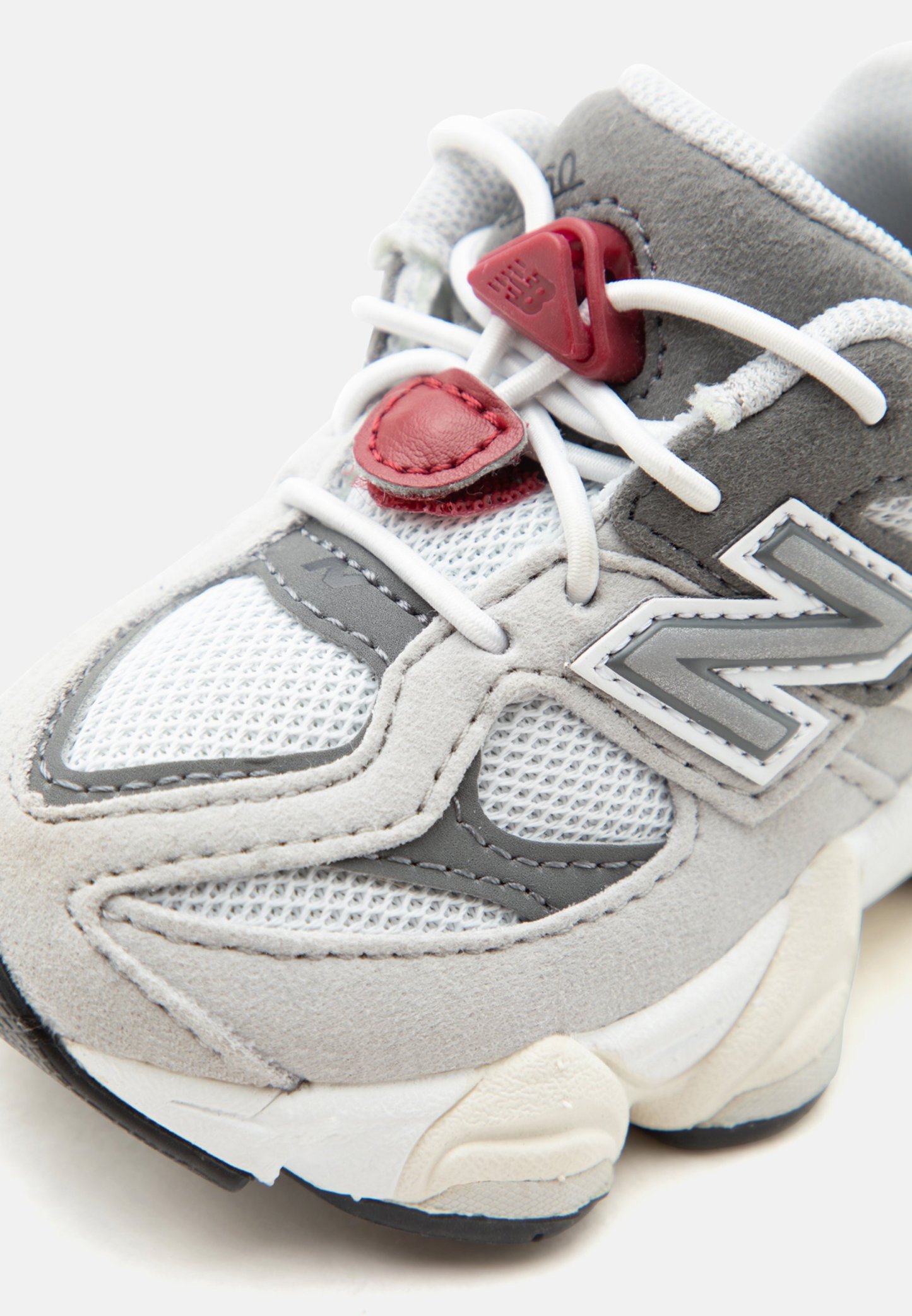 Zapatilla New Balance gris y blanca con paneles de malla y ante, cordones blancos y detalles de cierre de cordones rojos sobre un fondo blanco.