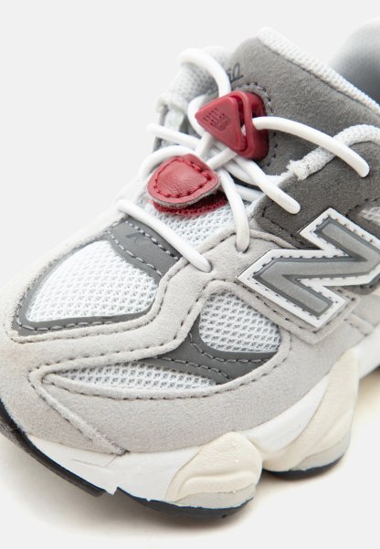 Zapatilla New Balance gris y blanca con paneles de malla y ante, cordones blancos y detalles de cierre de cordones rojos sobre un fondo blanco.