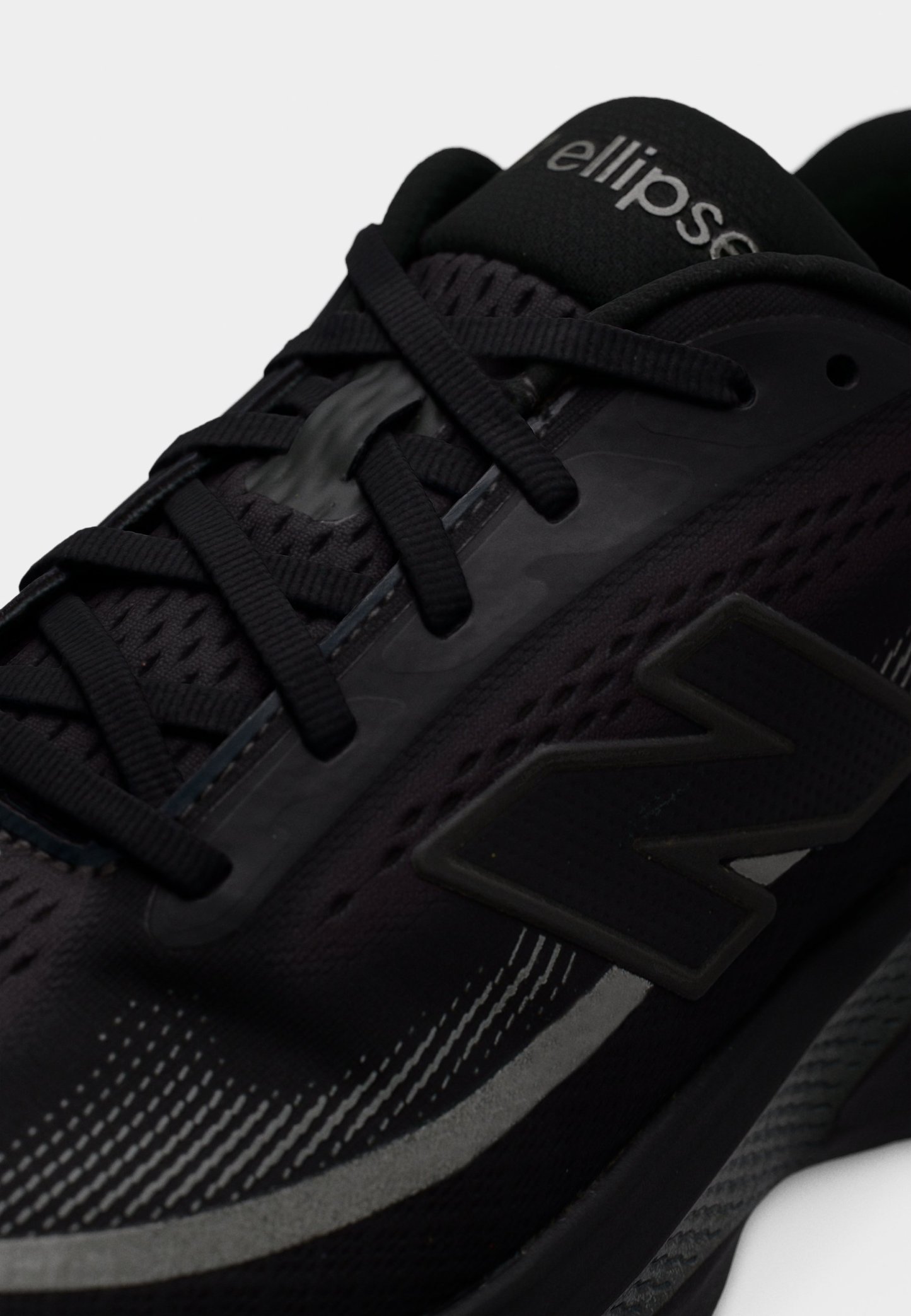 Zapatilla deportiva negra con parte superior de malla, detalles texturizados y elementos reflectantes. Cuenta con un logo gris prominente y cordones negros.