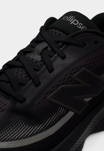 Zapatilla deportiva negra con parte superior de malla, detalles texturizados y elementos reflectantes. Cuenta con un logo gris prominente y cordones negros.