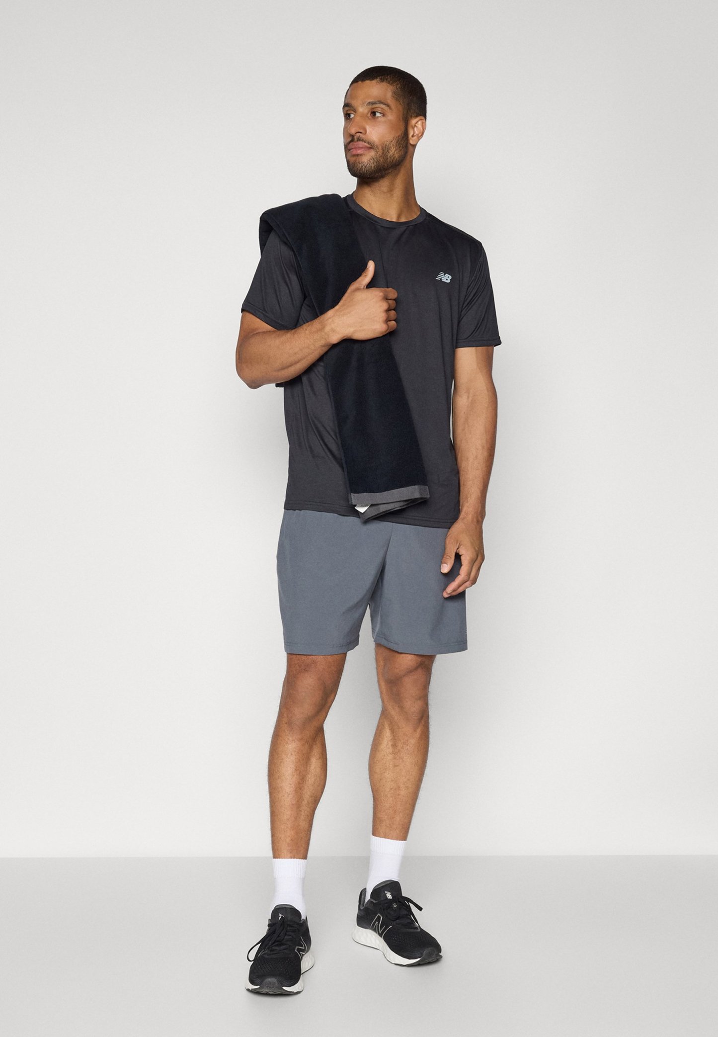 Hombre con camiseta deportiva negra, pantalones cortos grises y zapatillas negras sostiene una toalla oscura sobre su hombro, de pie contra un fondo sencillo.