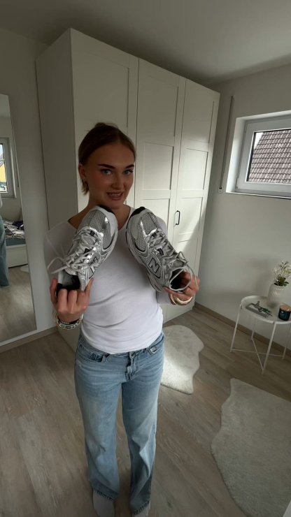 Zapatillas New Balance plateadas con detalles de malla y suelas negras, sostenidas por una persona con una camiseta blanca y jeans azul claro.