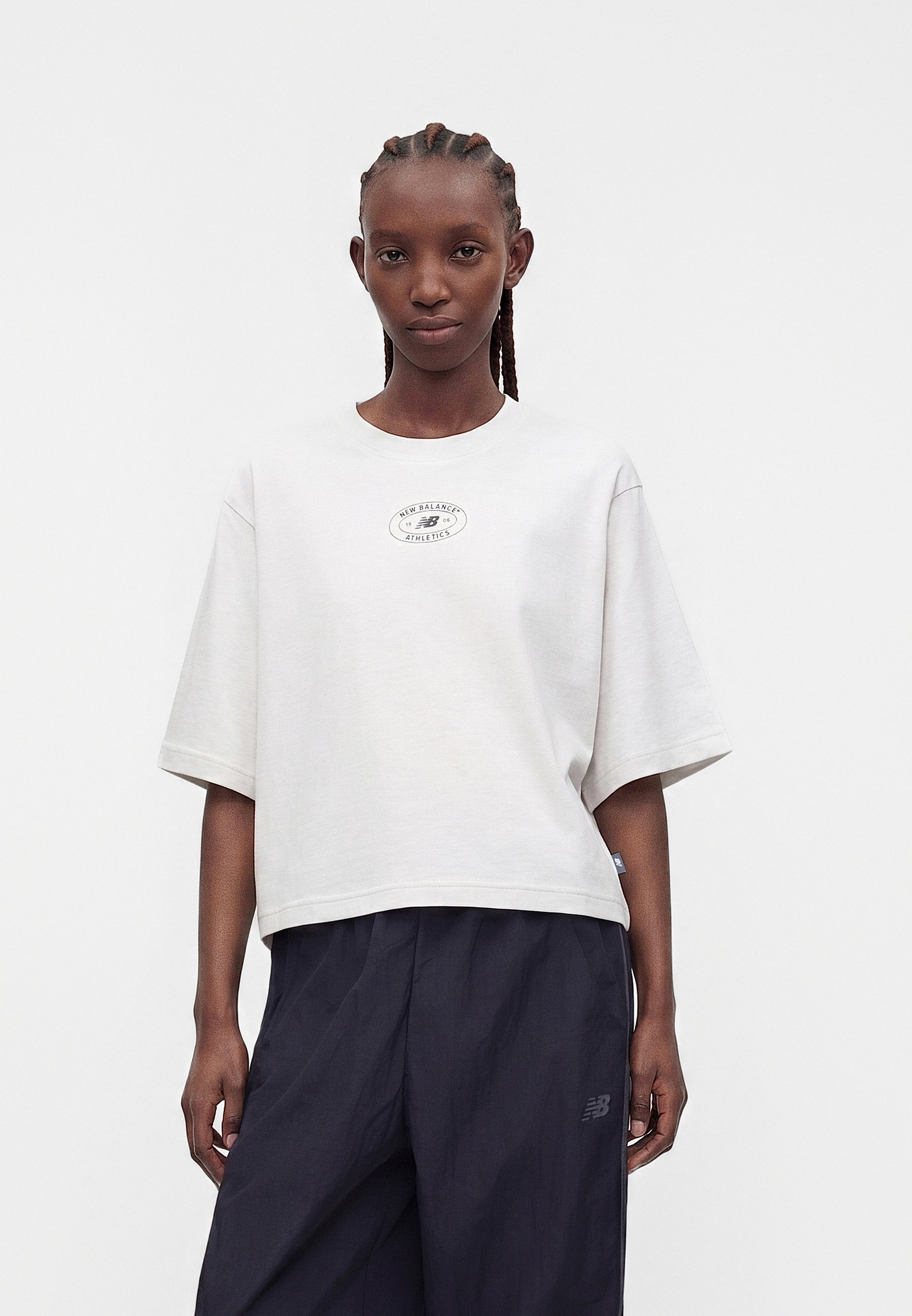 Mujer con el cabello trenzado que lleva una camiseta blanca oversize de New Balance y pantalones oscuros y sueltos, de pie contra un fondo claro y sencillo.