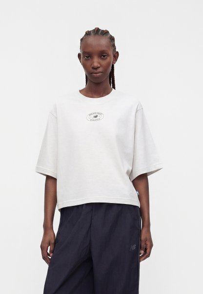 Mujer con el cabello trenzado que lleva una camiseta blanca oversize de New Balance y pantalones oscuros y sueltos, de pie contra un fondo claro y sencillo.