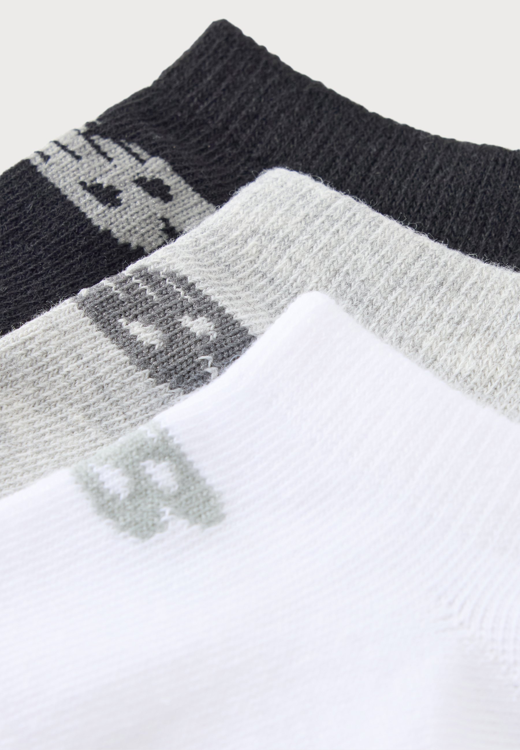 Tres pares de calcetines en negro, gris y blanco, cada uno con un pequeño logotipo geométrico cerca del puño, dispuestos en capas superpuestas.
