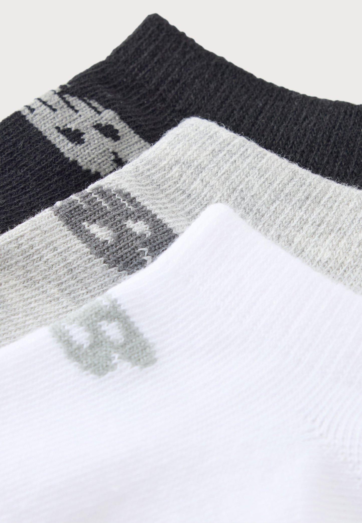 Tres pares de calcetines en negro, gris y blanco, cada uno con un pequeño logotipo geométrico cerca del puño, dispuestos en capas superpuestas.