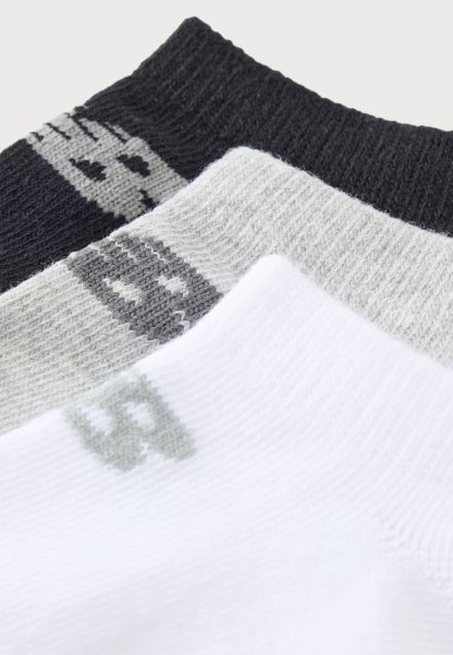 Tres pares de calcetines en negro, gris y blanco, cada uno con un pequeño logotipo geométrico cerca del puño, dispuestos en capas superpuestas.