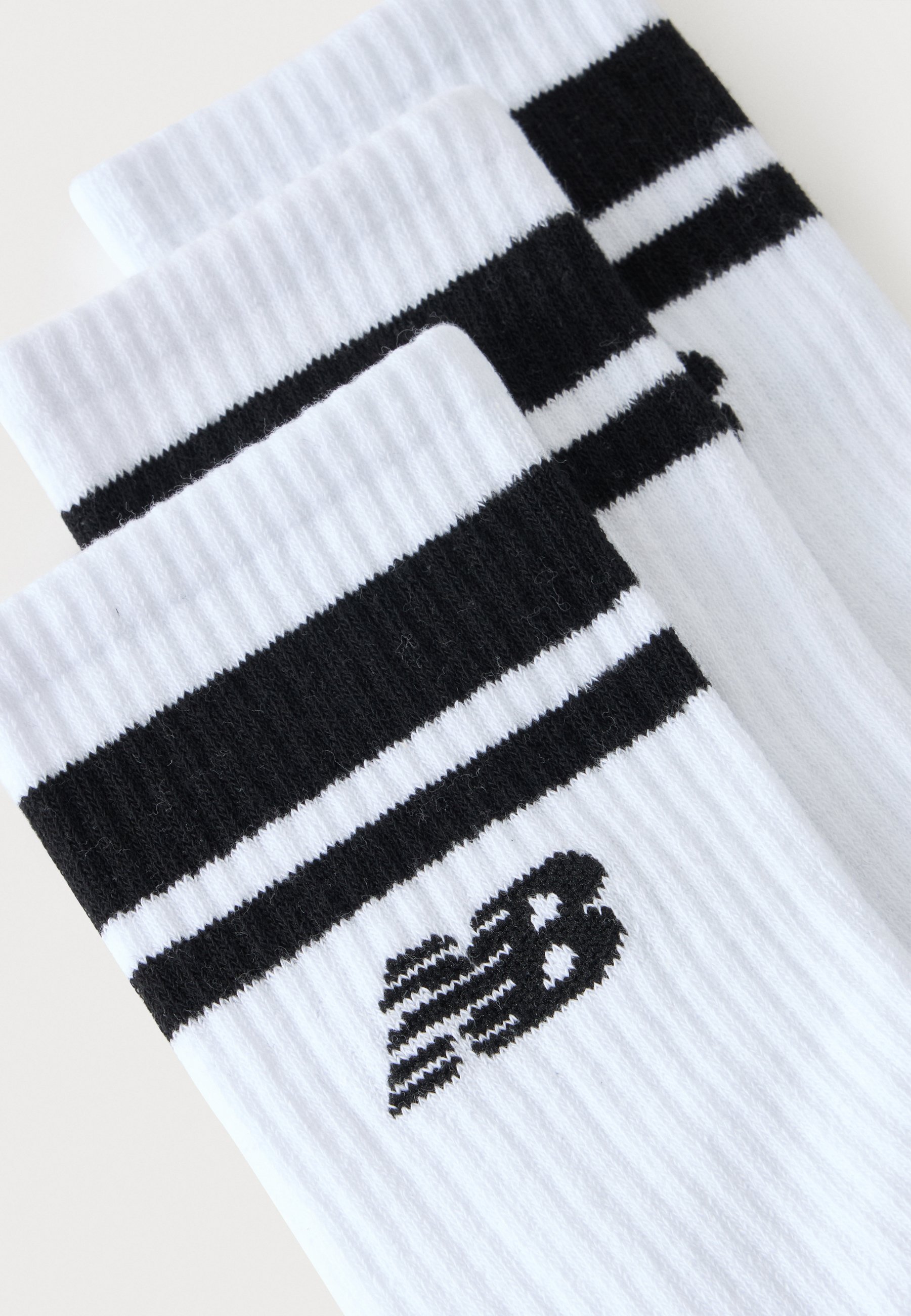 Tres calcetines blancos acanalados con rayas negras en la parte superior y un logo negro "NB" cerca del puño, apilados en diagonal.