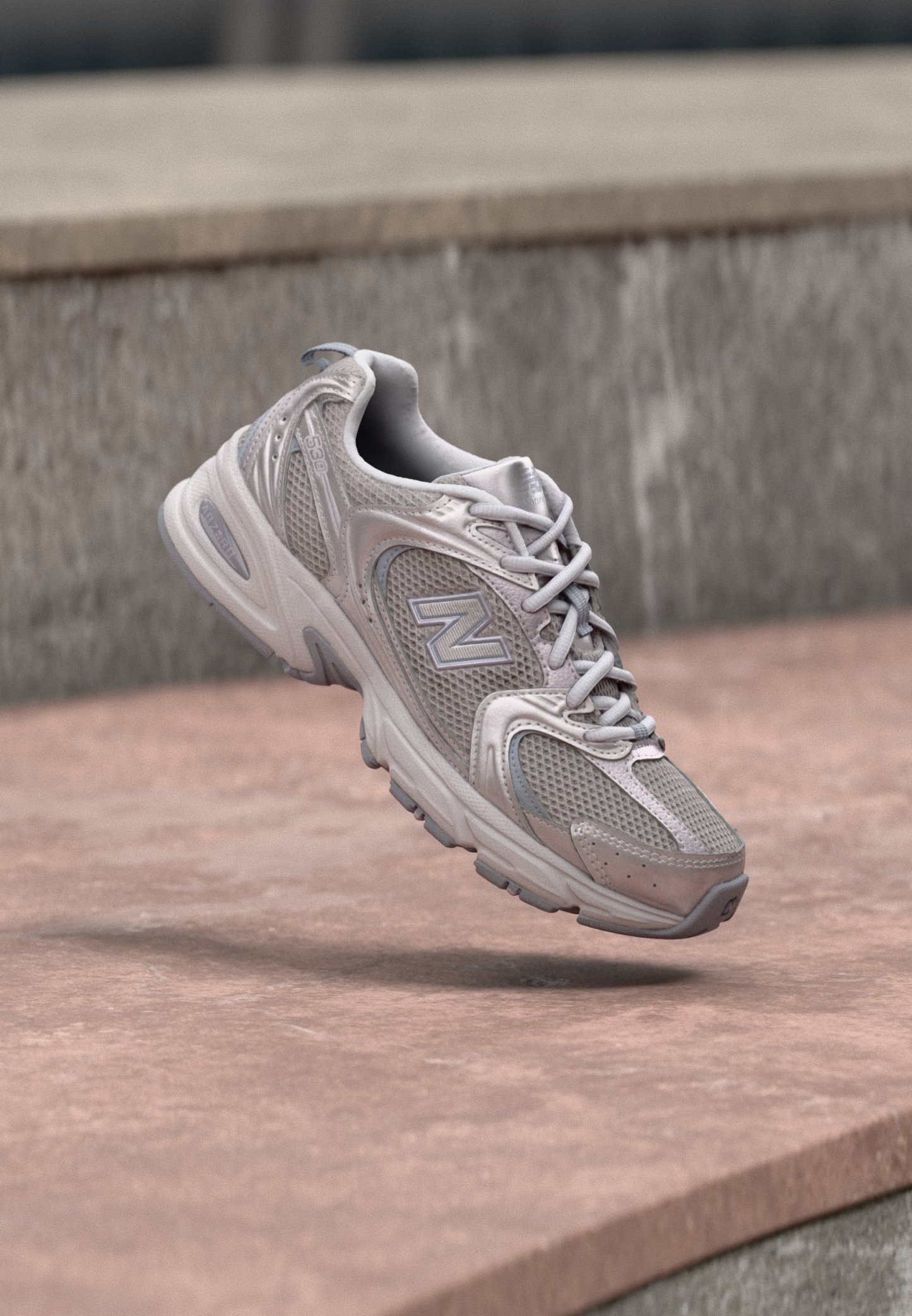 Zapatilla New Balance gris y blanca con detalles de malla y cuero, suspendida sobre una superficie de piedra con un fondo de pared de hormigón.