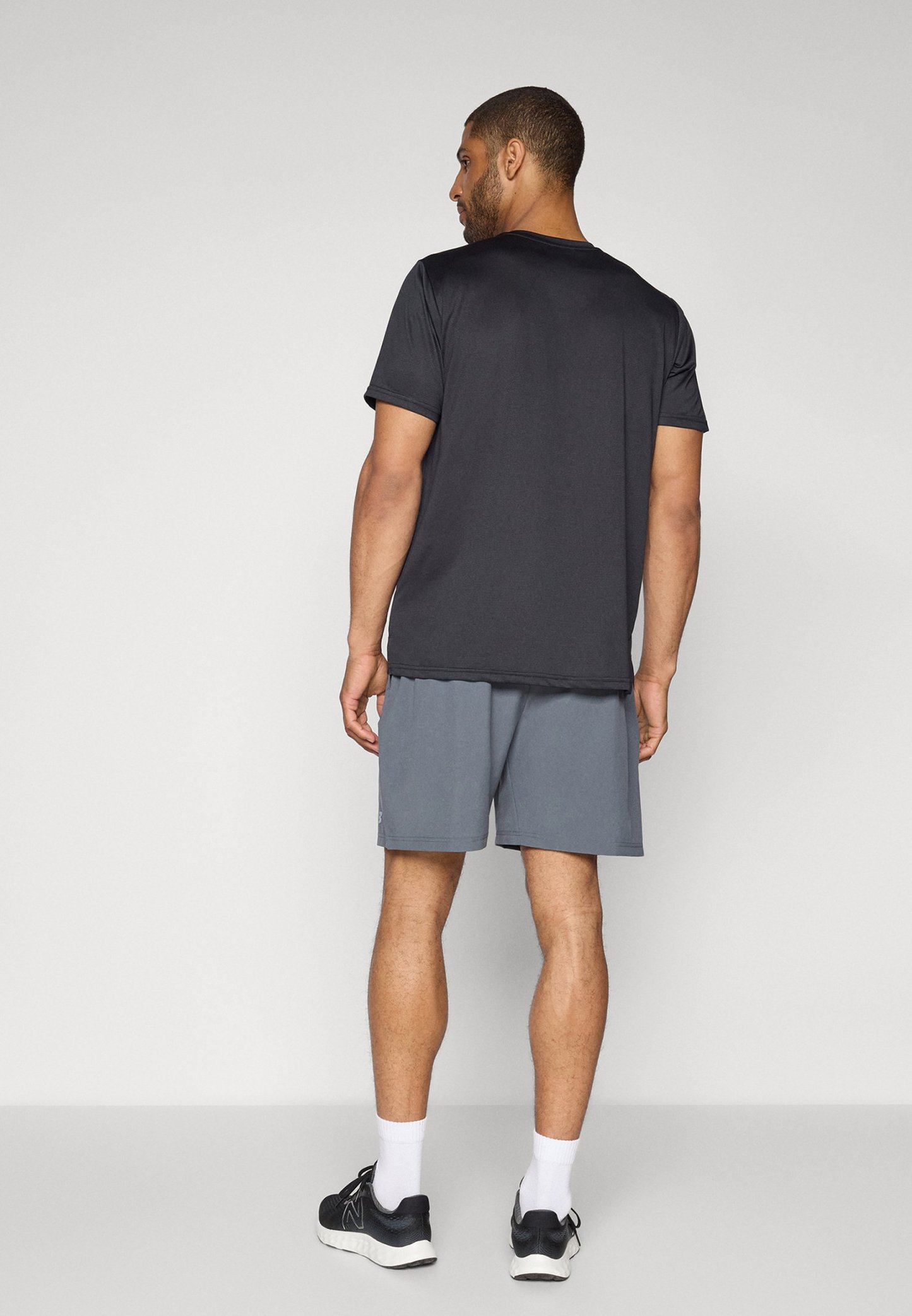 Hombre de pie de espaldas, vistiendo una camiseta negra de manga corta, pantalones cortos grises, calcetines blancos y zapatillas deportivas negras, frente a un fondo liso.