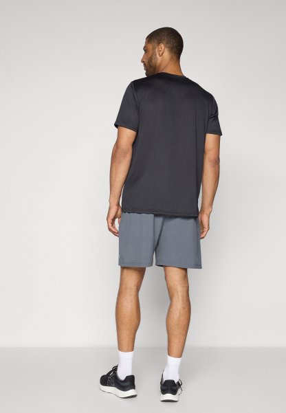 Hombre de pie de espaldas, vistiendo una camiseta negra de manga corta, pantalones cortos grises, calcetines blancos y zapatillas deportivas negras, frente a un fondo liso.