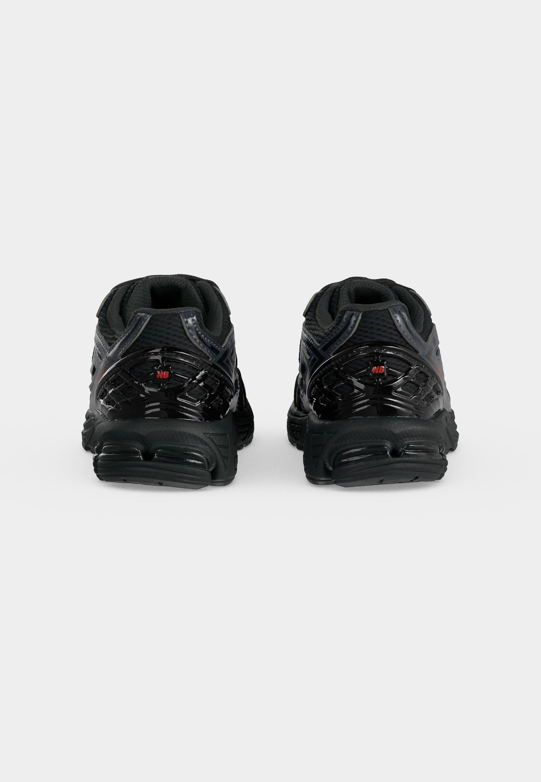Par de zapatillas deportivas negras vistas desde atrás, con suelas texturizadas y pequeños logotipos rojos "NB" en los contrafuertes del talón.