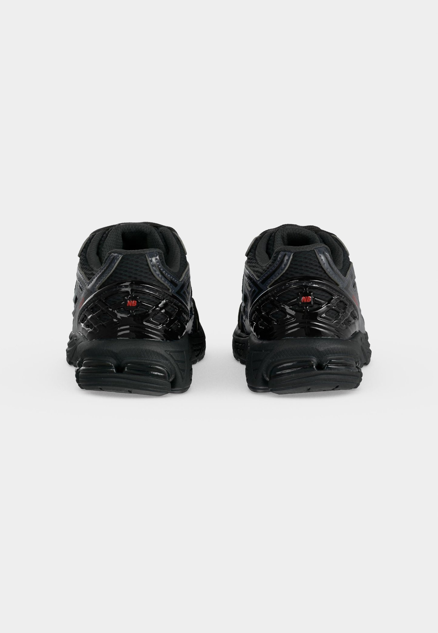 Par de zapatillas deportivas negras vistas desde atrás, con suelas texturizadas y pequeños logotipos rojos "NB" en los contrafuertes del talón.