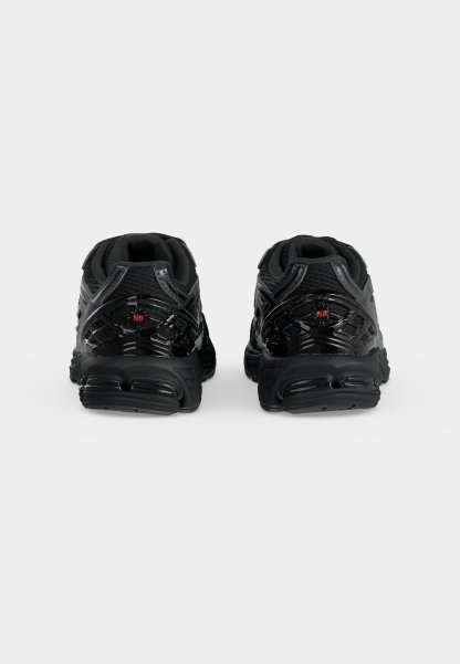 Par de zapatillas deportivas negras vistas desde atrás, con suelas texturizadas y pequeños logotipos rojos "NB" en los contrafuertes del talón.