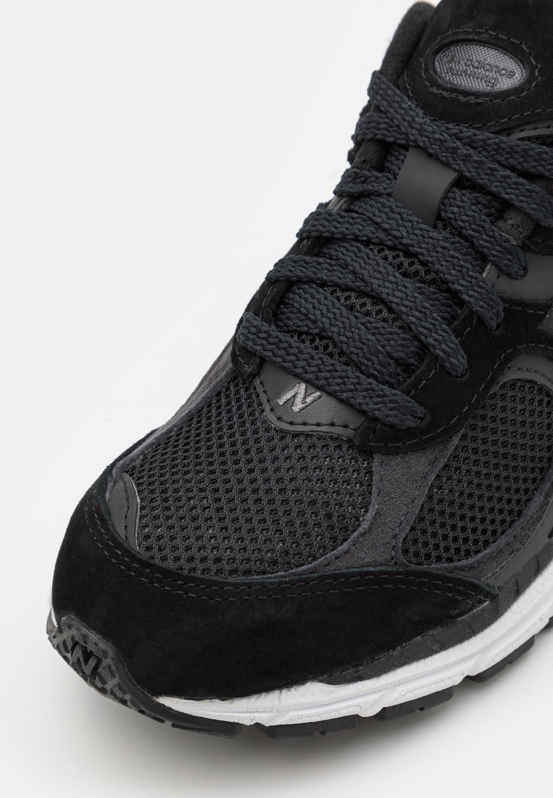 Zapatilla deportiva negra con parte superior de malla, detalles en ante, gruesos cordones negros y una entresuela blanca con detalles texturizados.