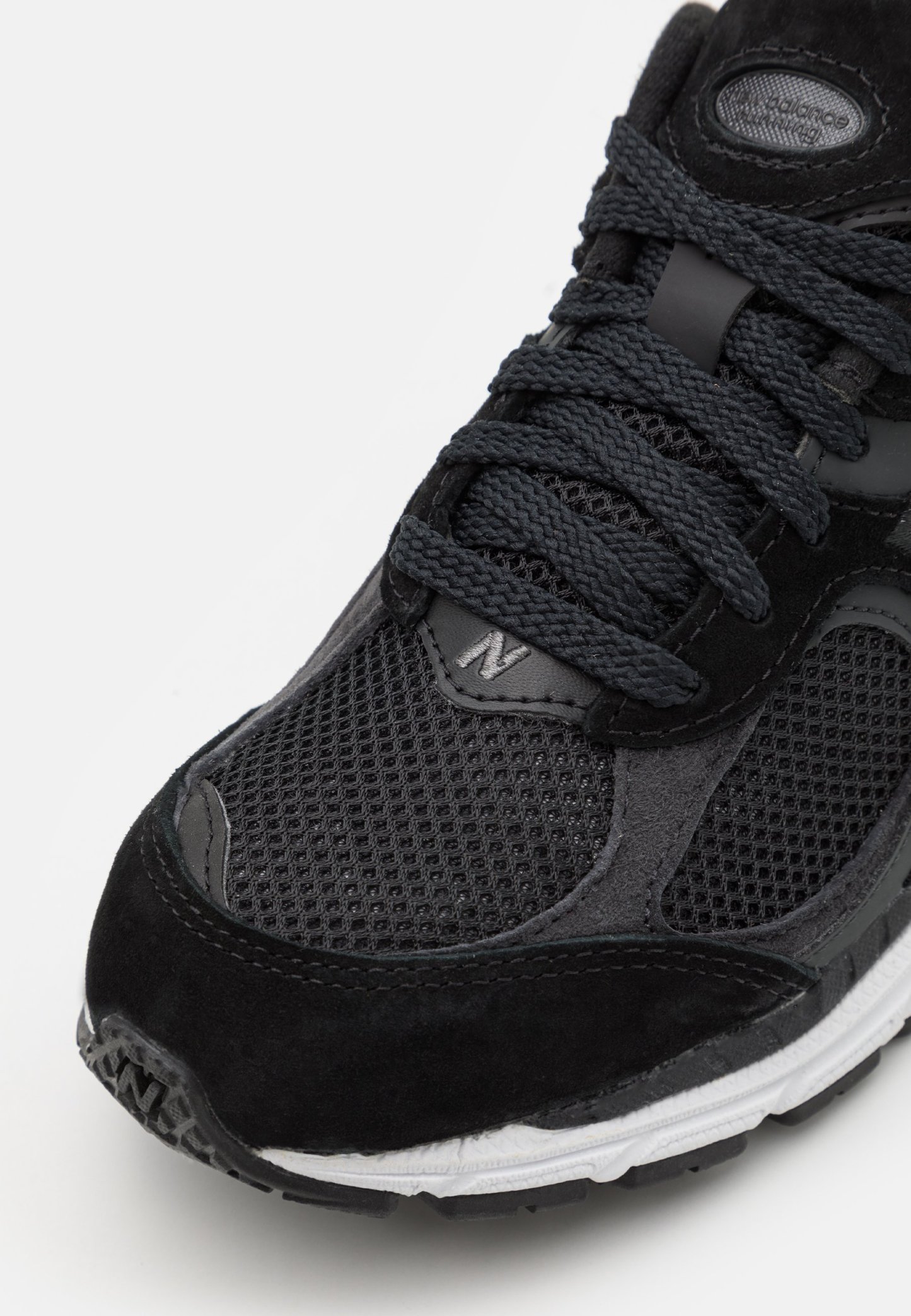 Zapatilla deportiva negra con parte superior de malla, detalles en ante, gruesos cordones negros y una entresuela blanca con detalles texturizados.