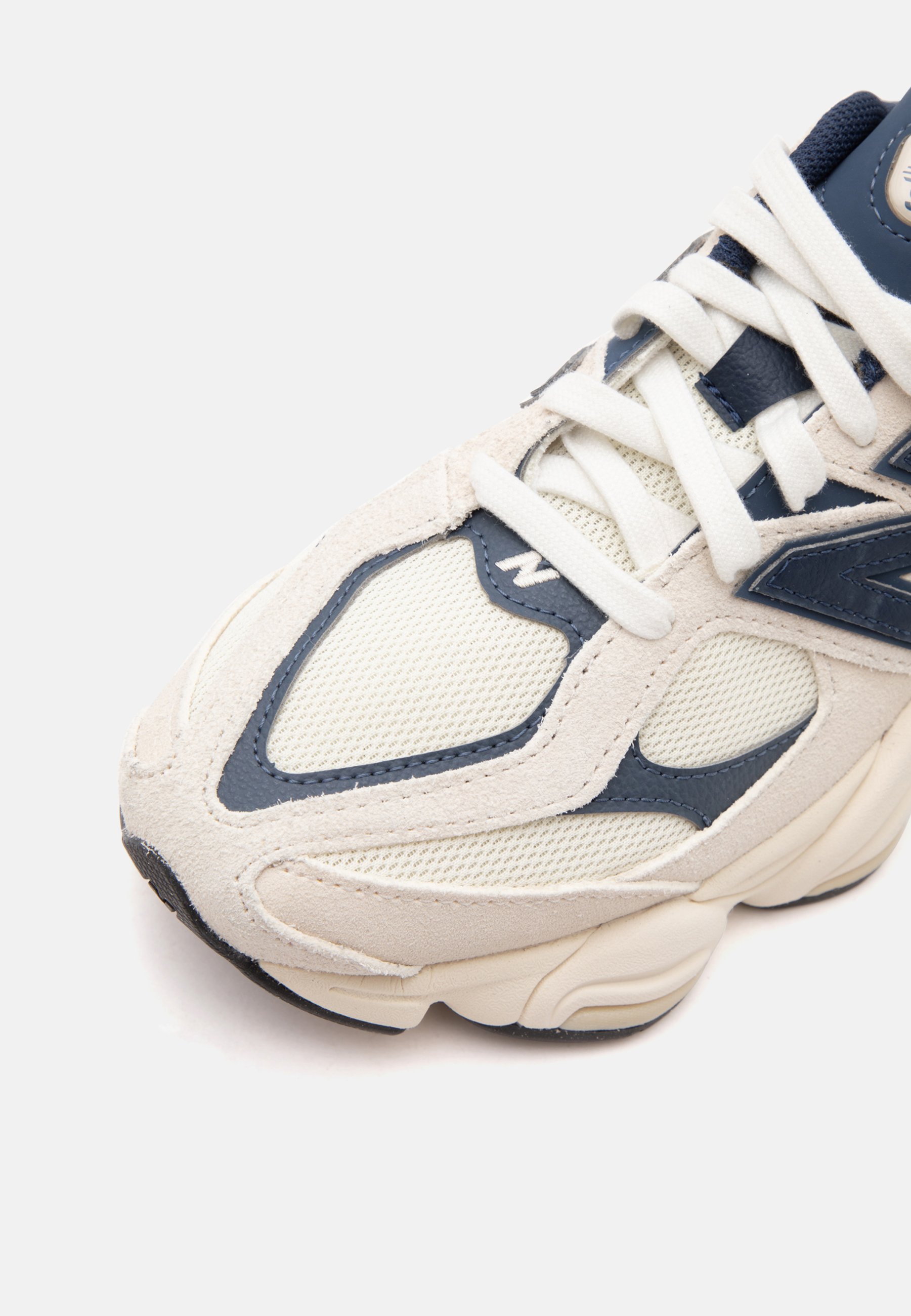 Zapatilla deportiva blanca y azul marino con texturas de malla y ante, enfocada en la parte delantera y los cordones sobre un fondo liso.