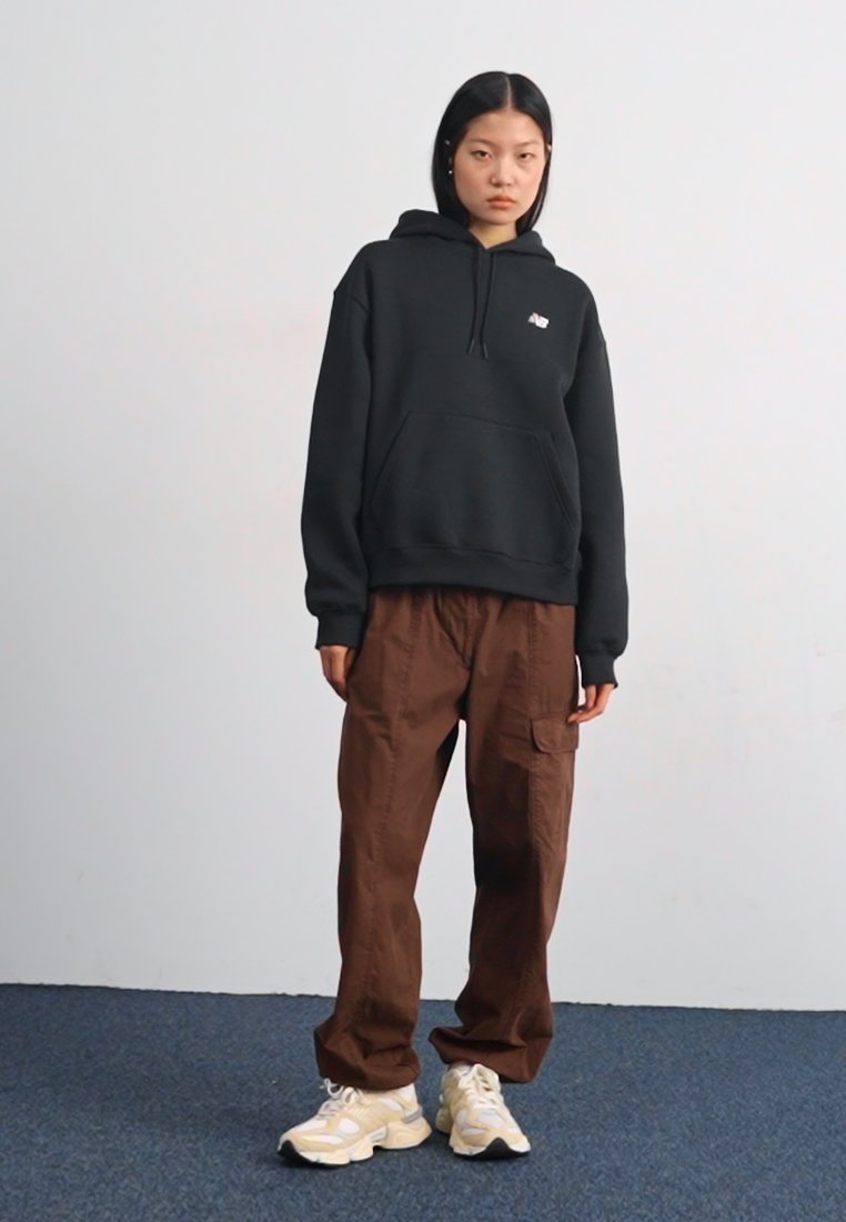 Sudadera con capucha negra y bolsillo frontal, combinada con pantalones cargo marrones y zapatillas de color crema. Tejido suave, corte relajado, logo visible.