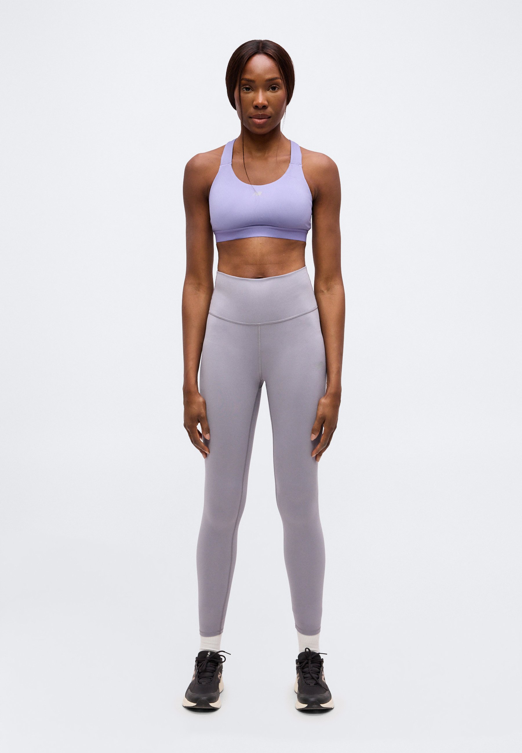 Mujer de pie, erguida, vestida con un sujetador deportivo lavanda, mallas grises de cintura alta, calcetines blancos y zapatillas negras sobre un fondo liso.