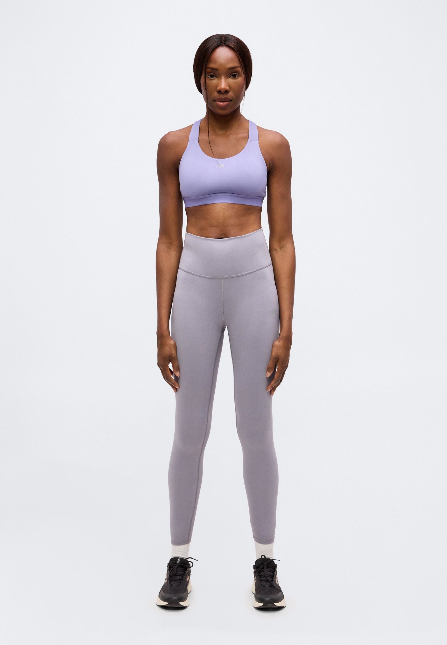Mujer de pie, erguida, vestida con un sujetador deportivo lavanda, mallas grises de cintura alta, calcetines blancos y zapatillas negras sobre un fondo liso.