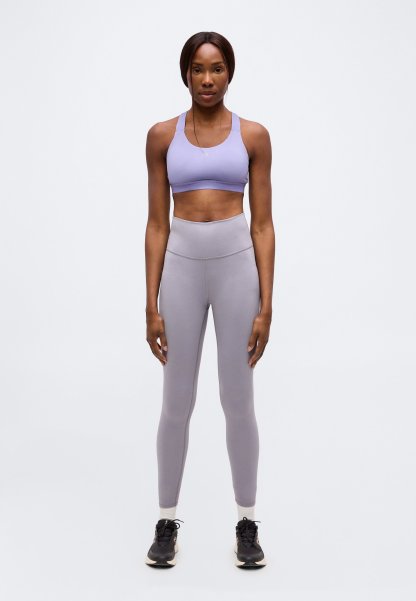 Mujer de pie, erguida, vestida con un sujetador deportivo lavanda, mallas grises de cintura alta, calcetines blancos y zapatillas negras sobre un fondo liso.