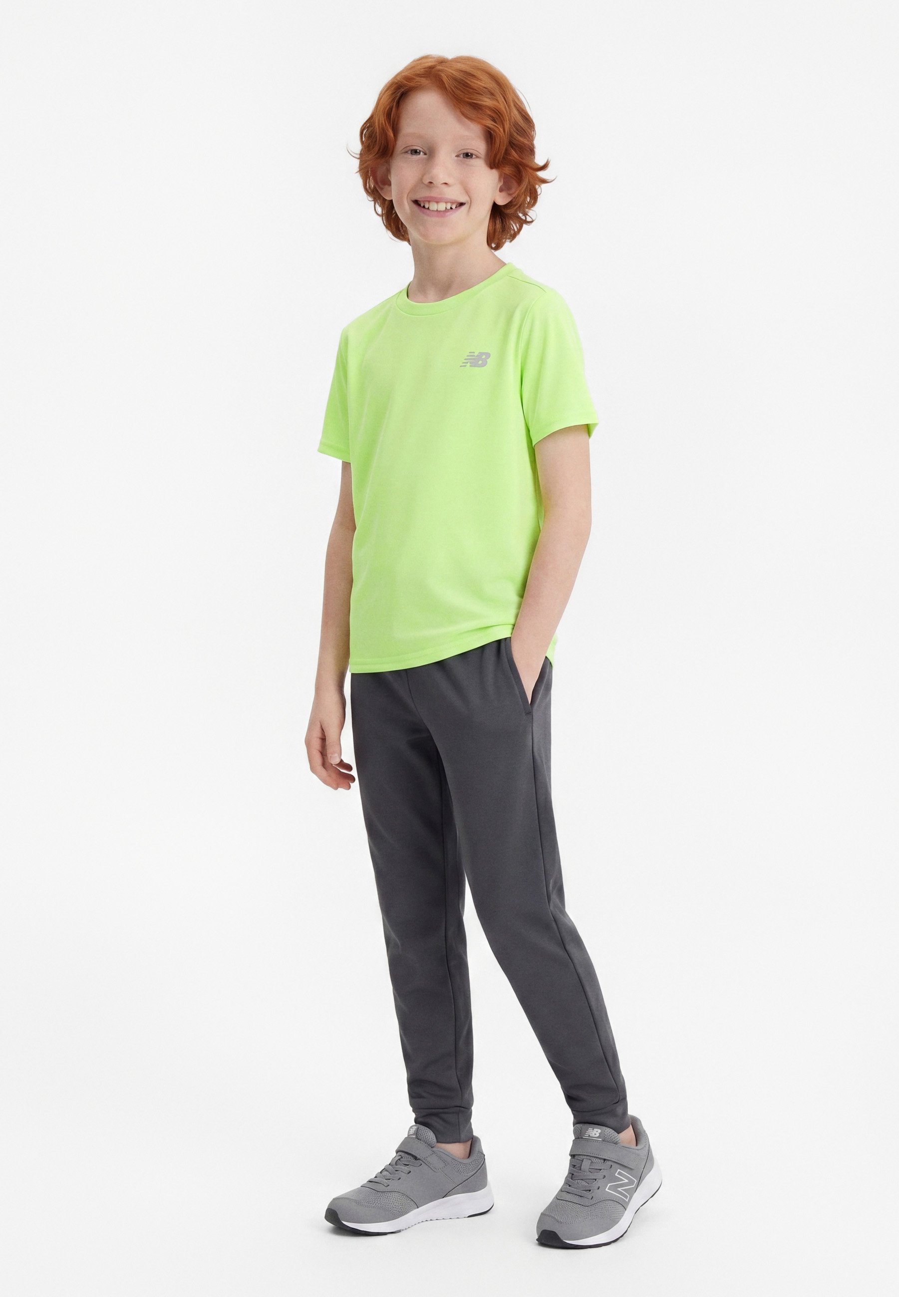 Niño pelirrojo sonriendo, de pie con las manos en los bolsillos, vestido con camiseta verde neón, pantalones gris oscuro y zapatillas grises sobre fondo blanco.