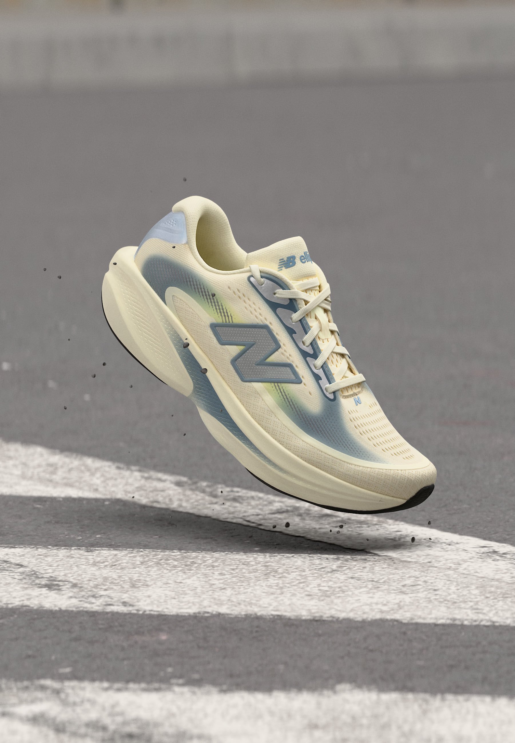 Zapatilla de running New Balance beige claro y azul en el aire sobre asfalto con marcas viales blancas y pequeños restos volando alrededor.