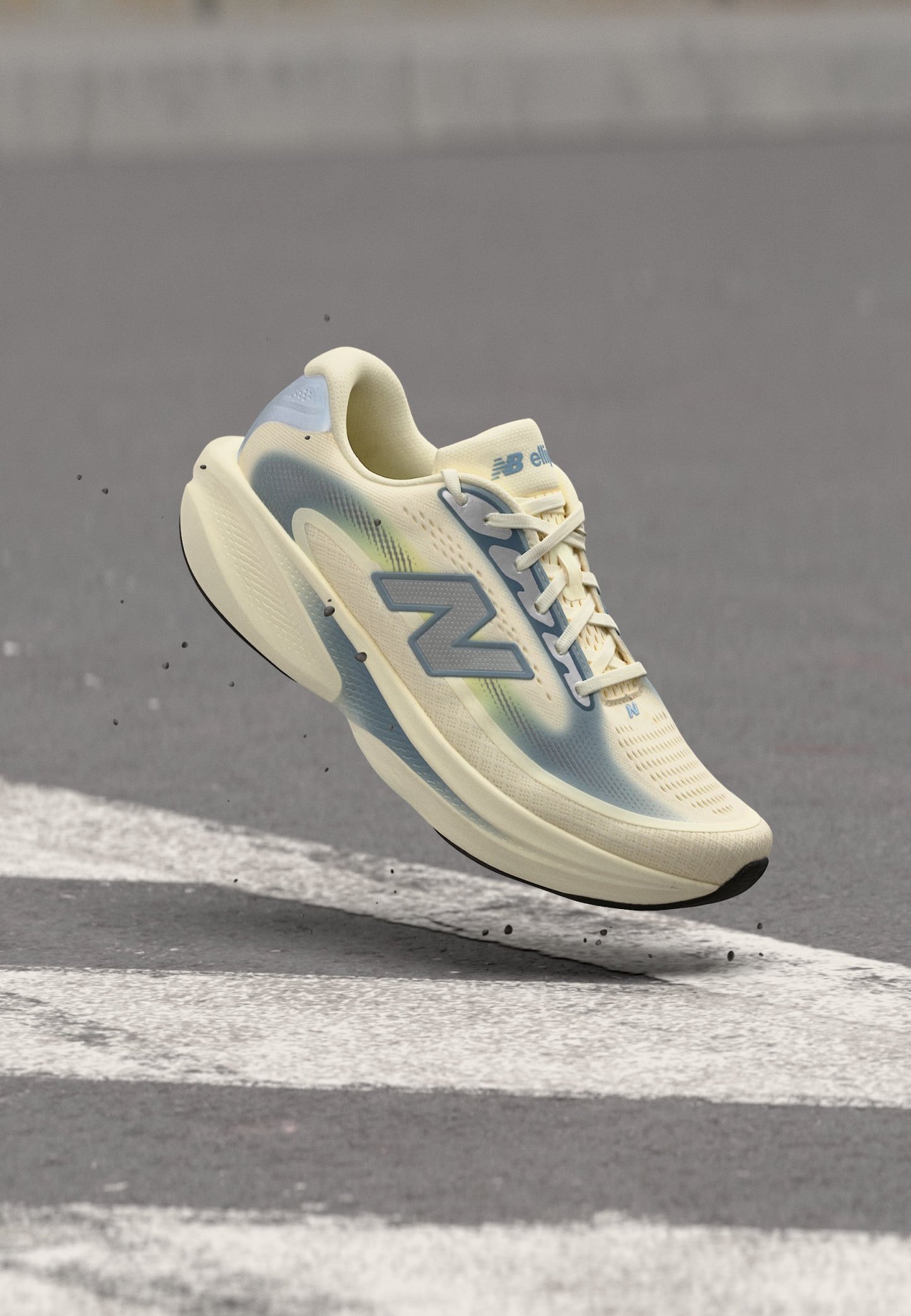 Zapatilla de running New Balance beige claro y azul en el aire sobre asfalto con marcas viales blancas y pequeños restos volando alrededor.