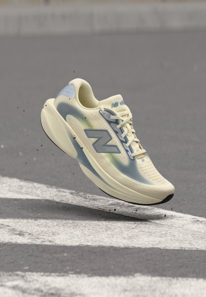 Zapatilla de running New Balance beige claro y azul en el aire sobre asfalto con marcas viales blancas y pequeños restos volando alrededor.