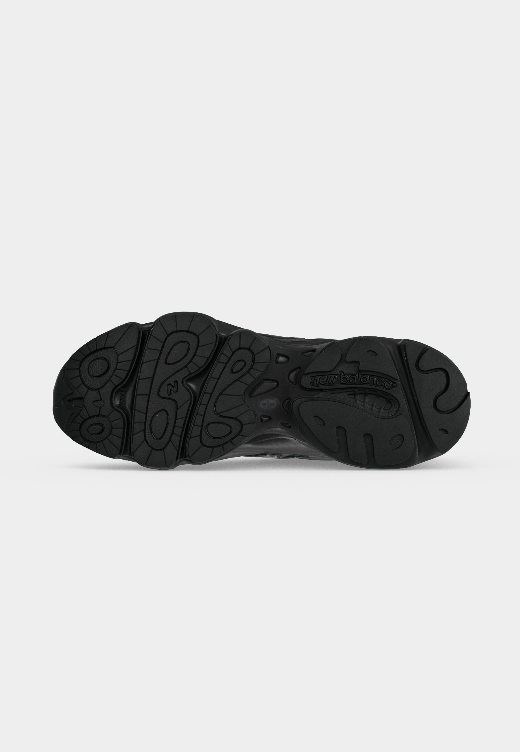 Suela negra de zapatilla New Balance con patrones texturizados antideslizantes y logo en relieve cerca del talón sobre un fondo gris claro liso.