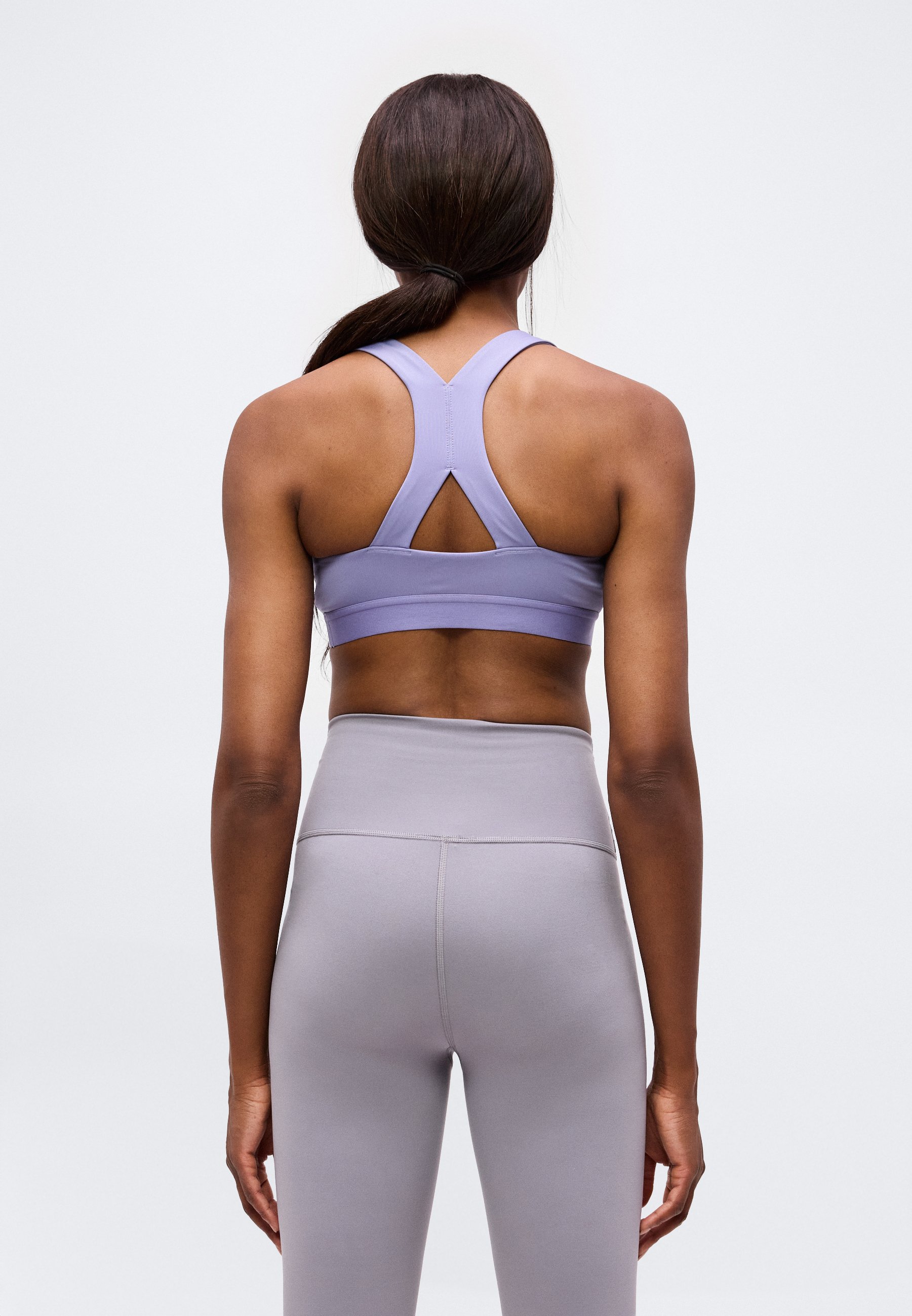 Mujer con cabello oscuro recogido en una coleta baja, vistiendo un sujetador deportivo lavanda y leggings gris claro de talle alto, vista desde atrás sobre un fondo liso.