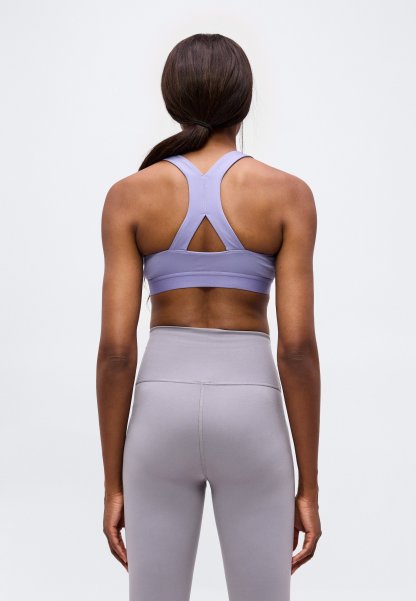 Mujer con cabello oscuro recogido en una coleta baja, vistiendo un sujetador deportivo lavanda y leggings gris claro de talle alto, vista desde atrás sobre un fondo liso.
