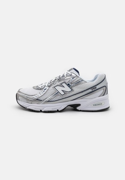 Zapatilla deportiva New Balance con parte superior de malla, predominantemente blanca con acentos en gris y azul marino, que presenta una suela acolchada y detalles texturizados.