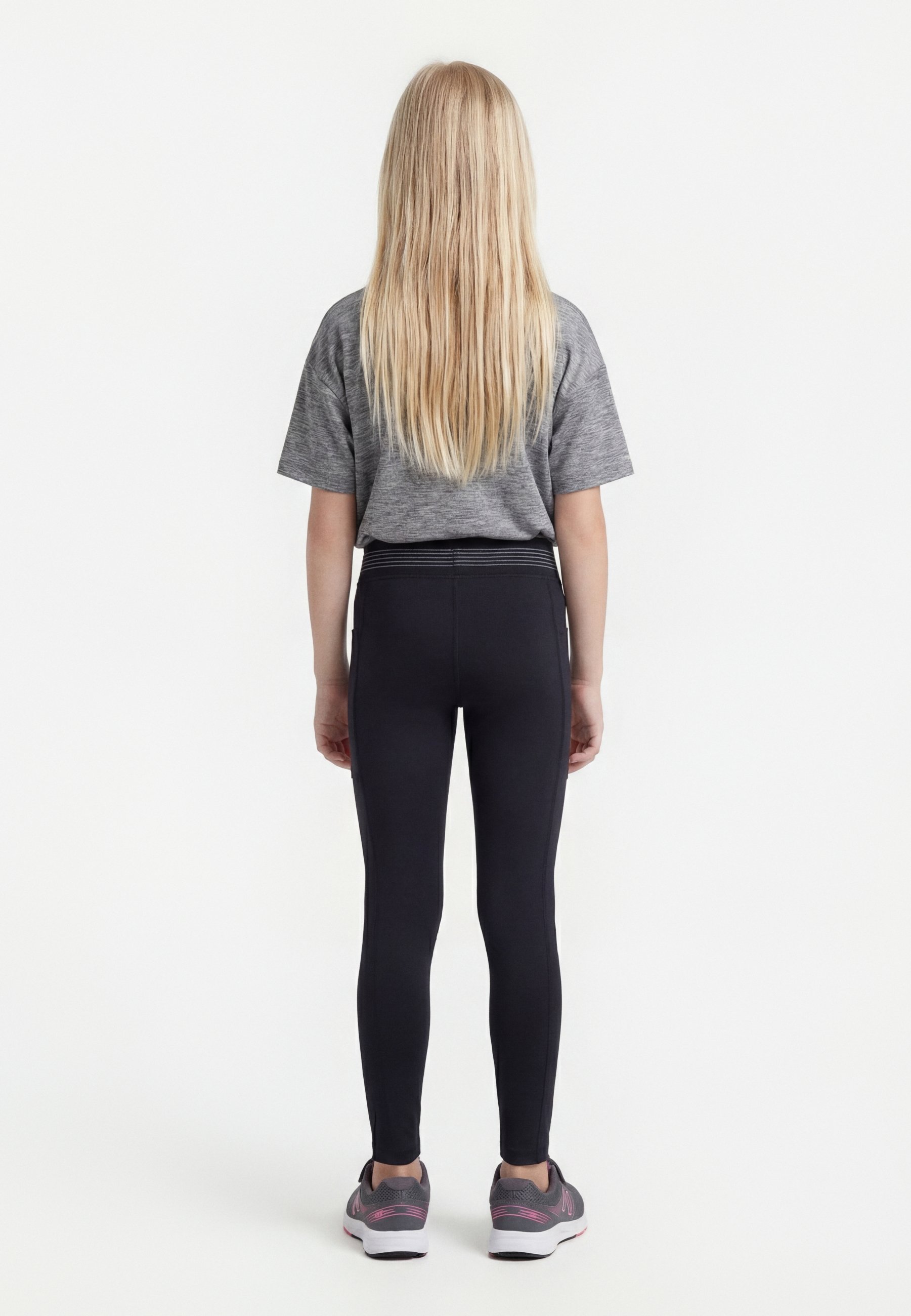 Niña rubia de pie de espaldas, con camiseta gris, mallas negras y zapatillas grises con detalles en rosa sobre un fondo blanco.